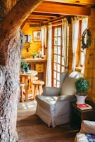 A cozy wooden interior featuring a large tree trunk integrated into the room's design. Sunlight streams through the glass-paned doors, illuminating a plush armchair with striped upholstery beside a wooden side table. Greenery in pots adds a fresh touch, and a small round wooden table with stools is visible near the wooden kitchenette.