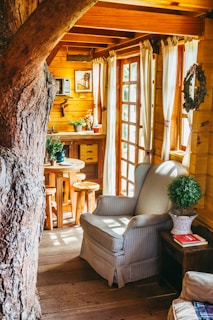 A cozy wooden interior featuring a large tree trunk integrated into the room's design. Sunlight streams through the glass-paned doors, illuminating a plush armchair with striped upholstery beside a wooden side table. Greenery in pots adds a fresh touch, and a small round wooden table with stools is visible near the wooden kitchenette.