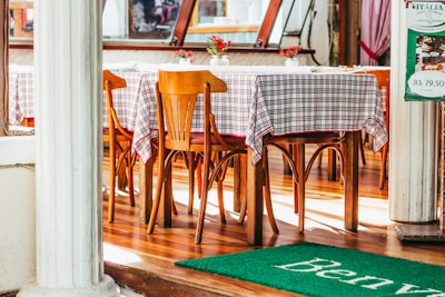 A cozy restaurant interior featuring wooden tables and chairs adorned with red and white checkered tablecloths. A decorative vase with red flowers is placed on each table, enhancing the warm and inviting atmosphere. The flooring is polished wood, and there are large windows allowing natural light to spill in. A green mat with partial lettering is placed at the entrance.