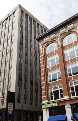 Two tall buildings with contrasting architectural styles, one modern and grey with many vertical windows and the other an older brick facade with arched windows and ornamental detailing. There is a street view below with a one-way sign and a partial view of a building's sign bearing the name 'Regions'. An American flag hangs on a pole.