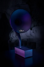 A modern, sleek phonograph-like speaker with a large, horn-shaped top emerging from a wooden base. The lighting casts a gradient of purples and blues, creating a futuristic and elegant appearance.