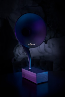 A modern, sleek phonograph-like speaker with a large, horn-shaped top emerging from a wooden base. The lighting casts a gradient of purples and blues, creating a futuristic and elegant appearance.