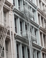 An architectural facade of a multi-story building featuring classic design elements like columns and decorative moldings. The exterior is painted in soft pastel tones, and several white metal fire escape ladders and platforms are visible running vertically and horizontally across the front.