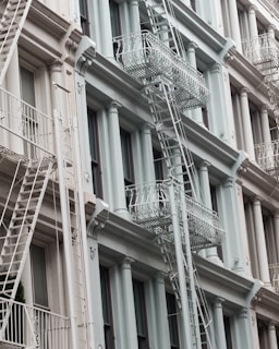 An architectural facade of a multi-story building featuring classic design elements like columns and decorative moldings. The exterior is painted in soft pastel tones, and several white metal fire escape ladders and platforms are visible running vertically and horizontally across the front.