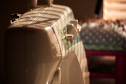 A sewing machine is partially visible, with a focus on its dials and a spool of thread. It is lit by warm light, creating a cozy and nostalgic atmosphere. In the background, a table with a patterned cloth is slightly blurred.
