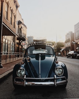 A vintage Beetle car is parked on a quiet street lined with old brick buildings. The sun is setting, casting a warm glow on the scene. The car has luggage strapped to its roof, suggesting a road trip or adventure.