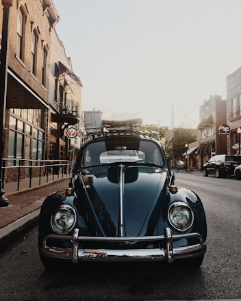 A vintage Beetle car is parked on a quiet street lined with old brick buildings. The sun is setting, casting a warm glow on the scene. The car has luggage strapped to its roof, suggesting a road trip or adventure.