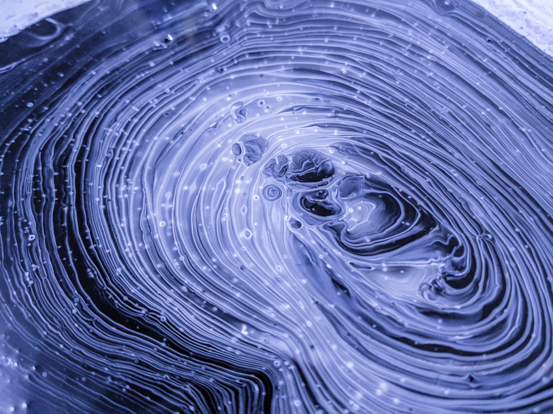 a close up of a water droplet with a blue background, This experimental painting features swirls of black, grey, purple, and white paints. As the paints began to dry, I took this closeup.<p></figure>The technique I use to create this kind of paint texture is called “fluid art,” and it relies on mixing a variety of mediums into acrylic paints to influence how they interact. I’ll use materials like glue, isopropyl alcohol, liquid silicone, and a butane torch.