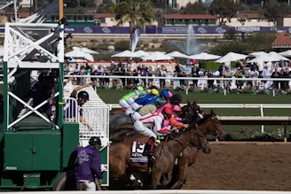 A group of jockeys on horseback are racing out of the starting gate on a dirt track. The horses and riders are adorned in colorful racing gear, and the scene is bustling with energy. In the background, a crowd of spectators watches from behind a fence, alongside tents and a fountain.