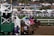 A group of jockeys on horseback are racing out of the starting gate on a dirt track. The horses and riders are adorned in colorful racing gear, and the scene is bustling with energy. In the background, a crowd of spectators watches from behind a fence, alongside tents and a fountain.