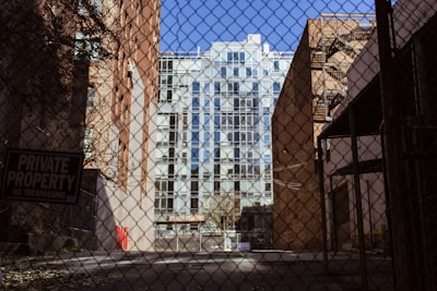 A chain-link fence in the foreground with a 'Private Property No Trespassing' sign. Beyond the fence, there is a narrow alleyway flanked by tall brick buildings. In the background, a modern building with glass windows is visible. The setting appears urban and slightly deserted.