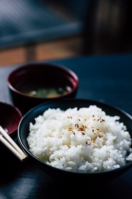 A bowl filled with steaming white rice garnished with sesame seeds is the main focus. Next to it, a pair of chopsticks rests on the table. In the background, a red bowl, possibly containing soup, is slightly out of focus. The setup suggests a traditional Japanese meal with a minimalist aesthetic.