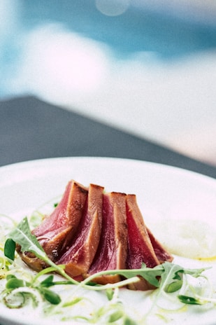 Thinly sliced seared tuna is arranged elegantly on a white plate, garnished with fresh greens. The tuna appears pink and lightly charred at the edges, with a glossy surface. The background is softly blurred, providing a contrast to the vibrant colors of the dish.