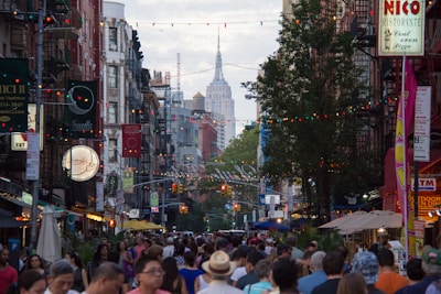 A bustling city street crowded with people, adorned with colorful string lights. Tall buildings line the street, with the Empire State Building visible in the background. Various restaurant signs and storefronts are lit up, adding to the vibrant atmosphere.