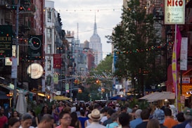 A bustling city street crowded with people, adorned with colorful string lights. Tall buildings line the street, with the Empire State Building visible in the background. Various restaurant signs and storefronts are lit up, adding to the vibrant atmosphere.