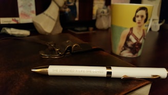 A white pen with gold accents lies on a dark leather-bound notebook. The pen is inscribed with the words 'SHE DESIGNED A LIFE SHE LOVED'. In the background, there is a colorful mug with an image of a woman printed on it. The setting appears to be a desk with various blurred objects in the background.
