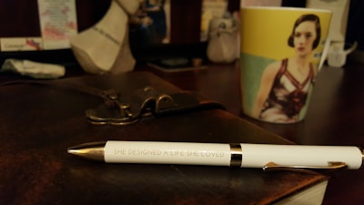 A white pen with gold accents lies on a dark leather-bound notebook. The pen is inscribed with the words 'SHE DESIGNED A LIFE SHE LOVED'. In the background, there is a colorful mug with an image of a woman printed on it. The setting appears to be a desk with various blurred objects in the background.