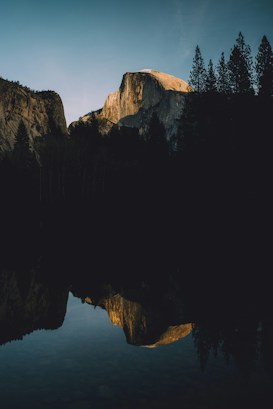 A landscape with a towering granite formation bathed in the golden light of sunset, standing above a calm, reflective surface of water. Dense pine trees frame the scene, adding depth to the silhouette of the horizon.