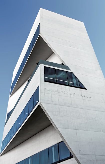 A modern architectural building with sharp, angular lines and large glass windows. The structure features multiple layers and clean, minimalist design, accented with the inscription 'Fondazione Prada' on its facade. The sky serves as a clear blue backdrop.