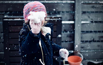A child wearing a colorful knitted hat and dark jacket is holding a snowball, with snowflakes falling around. The background features a wooden fence and a red bucket.