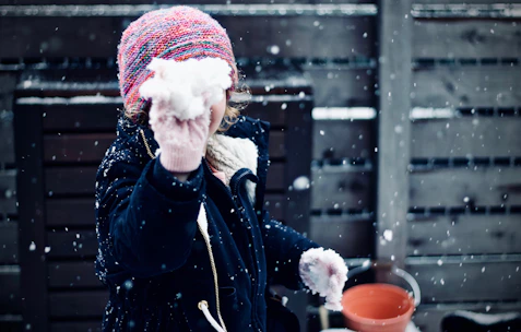 A child wearing a colorful knitted hat and dark jacket is holding a snowball, with snowflakes falling around. The background features a wooden fence and a red bucket.