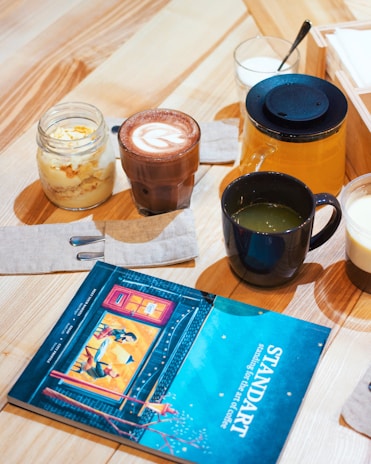 A wooden table is set with a variety of beverages and a dessert. There is a glass of hot chocolate with latte art, a glass jar containing a dessert, and a black cup filled with a green beverage. Next to these items, there is a glass pitcher with a dark blue lid, likely containing juice or tea, and an open magazine with a colorful cover featuring an illustration. The table is also adorned with napkins and a neatly arranged spoon.