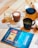 A wooden table is set with a variety of beverages and a dessert. There is a glass of hot chocolate with latte art, a glass jar containing a dessert, and a black cup filled with a green beverage. Next to these items, there is a glass pitcher with a dark blue lid, likely containing juice or tea, and an open magazine with a colorful cover featuring an illustration. The table is also adorned with napkins and a neatly arranged spoon.