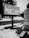 A black and white photograph featuring a sign reading 'Happiness Inn' at Niagara Falls, with words 'Secluded' and 'Serene' also visible. The area is covered in snow with patches of grass, and a 'Vacancy' sign lies on the ground near a paved road.