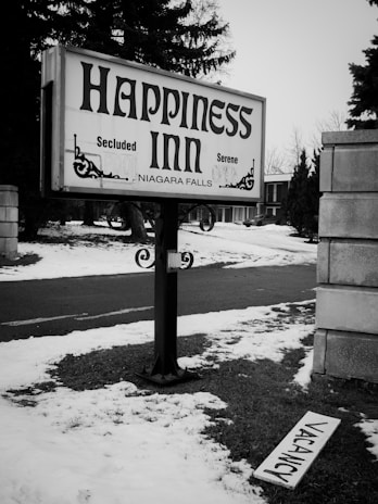 A black and white photograph featuring a sign reading 'Happiness Inn' at Niagara Falls, with words 'Secluded' and 'Serene' also visible. The area is covered in snow with patches of grass, and a 'Vacancy' sign lies on the ground near a paved road.