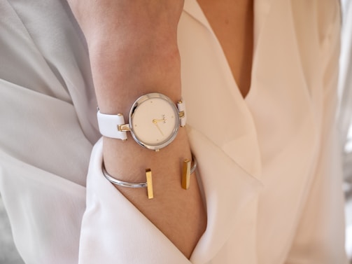 A person wearing a white watch on the wrist, complemented by a metallic bracelet with gold accents. The person is dressed in a light, cream-colored garment, possibly a shirt or blouse. The watch has a minimalistic design with gold markers and a sleek, white strap.