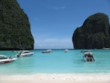 Crystal-clear turquoise waters gently lap against a pristine sandy beach surrounded by towering limestone cliffs covered in green foliage. Several boats, including speedboats and traditional long-tail boats, are anchored or moving across the water, hinting at leisure or tour activities. The sky is a bright blue with a few wispy clouds.