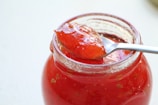 A clear glass jar filled with vibrant red jam is featured, with a shiny metal spoon resting on top. The spoon holds a generous portion of the jam, showcasing its glossy and rich texture. The jam appears to have chunks of fruit, indicating a homemade or artisanal quality.