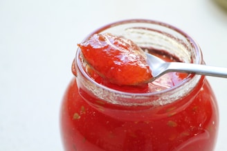 A clear glass jar filled with vibrant red jam is featured, with a shiny metal spoon resting on top. The spoon holds a generous portion of the jam, showcasing its glossy and rich texture. The jam appears to have chunks of fruit, indicating a homemade or artisanal quality.
