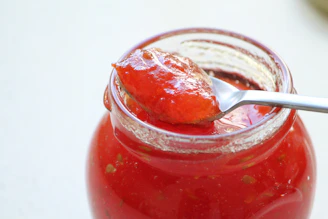 A rustic wooden table topped with a variety of vibrant homemade jam jars, fresh fruit, and a spoon drizzling golden jelly.