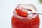A clear glass jar filled with vibrant red jam is featured, with a shiny metal spoon resting on top. The spoon holds a generous portion of the jam, showcasing its glossy and rich texture. The jam appears to have chunks of fruit, indicating a homemade or artisanal quality.