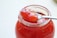 A clear glass jar filled with vibrant red jam is featured, with a shiny metal spoon resting on top. The spoon holds a generous portion of the jam, showcasing its glossy and rich texture. The jam appears to have chunks of fruit, indicating a homemade or artisanal quality.