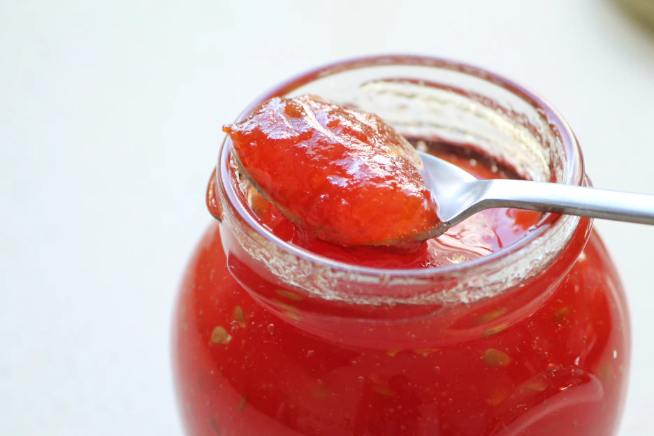 A rustic wooden table topped with a variety of vibrant homemade jam jars, fresh fruit, and a spoon drizzling golden jelly.