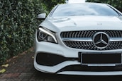 A luxury car with a sleek design, featuring a prominent grille and distinctive headlight. The vehicle is parked on a brick driveway, surrounded by lush greenery on one side.