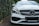 A luxury car with a sleek design, featuring a prominent grille and distinctive headlight. The vehicle is parked on a brick driveway, surrounded by lush greenery on one side.