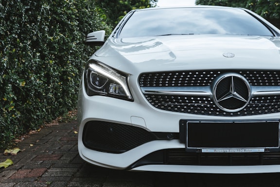 A luxury car with a sleek design, featuring a prominent grille and distinctive headlight. The vehicle is parked on a brick driveway, surrounded by lush greenery on one side.