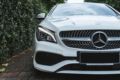 A luxury car with a sleek design, featuring a prominent grille and distinctive headlight. The vehicle is parked on a brick driveway, surrounded by lush greenery on one side.