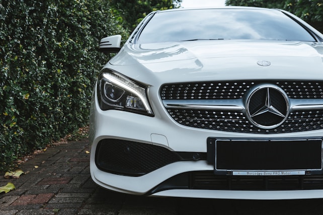 A luxury car with a sleek design, featuring a prominent grille and distinctive headlight. The vehicle is parked on a brick driveway, surrounded by lush greenery on one side.