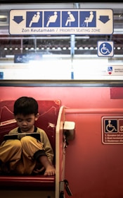 A young child sits alone in a priority seating area on public transport, engrossed in a handheld device. The seating is marked by signage indicating special priority for certain passengers. The overall setting seems quiet and focused.
