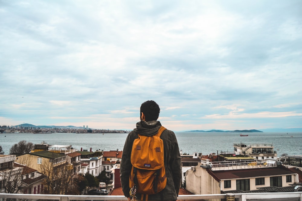 man carrying orange backpack looking at the houses near ocean