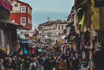 A bustling outdoor market scene with numerous people walking between stalls on either side. The market is filled with a variety of colorful textiles and clothing hanging from canopies. Buildings in the background show mixed architectural styles, some with signs in a foreign language. The atmosphere seems lively and vibrant, indicative of a busy and popular shopping area.