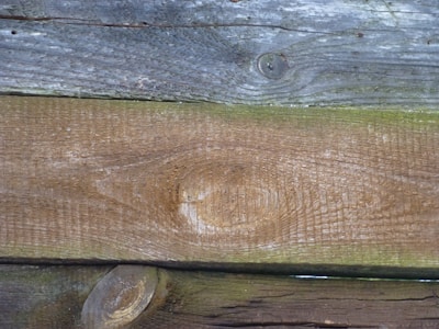 Weathered wooden planks with distinct grain patterns and knots, displaying varying shades of brown and grey. The wood appears worn and aged, indicating exposure to the elements.