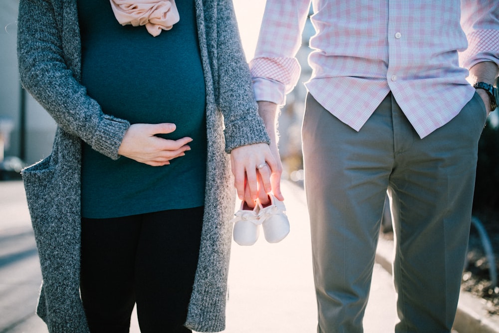 man standing beside pregnant woman holding baby´s shoes during daytime