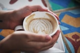 A cup of coffee is being held by two hands, with latte art on the surface resembling a leaf pattern. The background features a colorful, geometric pattern on a tiled surface.