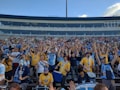 A large group of enthusiastic sports fans, many wearing team jerseys in yellow and blue, are gathered in a stadium. They have their hands raised, showing excitement and support. Some fans hold scarves, while one person near the front is wearing a luchador mask. A few drums are visible, contributing to the lively atmosphere.
