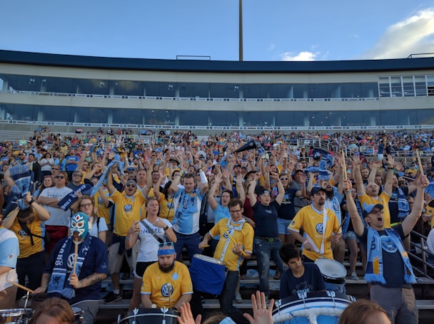 A large group of enthusiastic sports fans, many wearing team jerseys in yellow and blue, are gathered in a stadium. They have their hands raised, showing excitement and support. Some fans hold scarves, while one person near the front is wearing a luchador mask. A few drums are visible, contributing to the lively atmosphere.